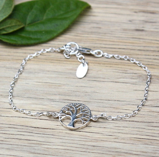 il y a un bracelet femme réalisé avec une chaine associé à un arbre de vie en argent 925 présenté sur une planche de bois avec de la verdure